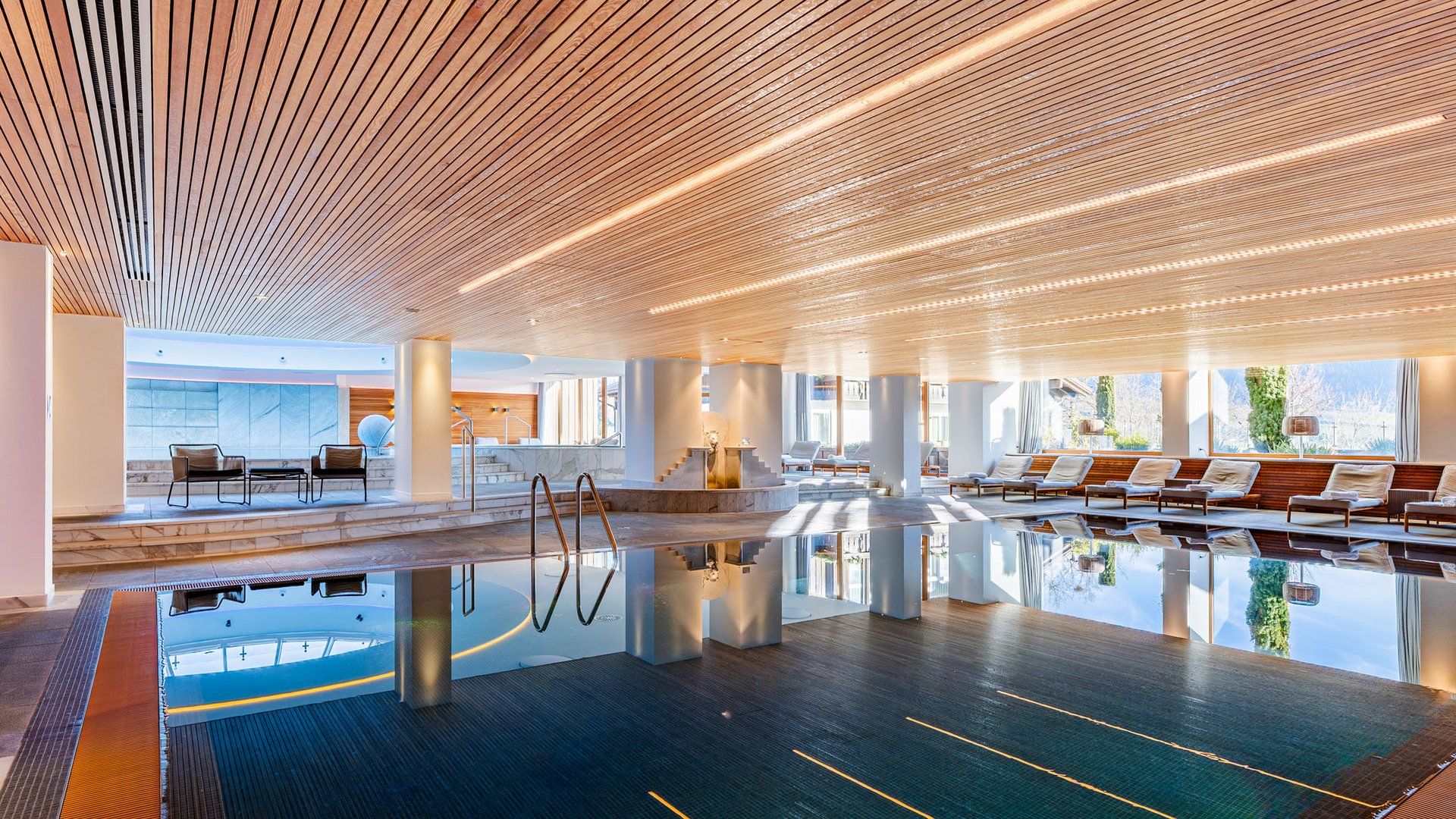 Innenpoolbereich mit Holzdecke, Liegestühlen und großen Fenstern mit Bergblick