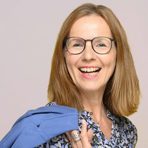 Lächelnde Frau mit Brille, Blumenbluse und blauem Pullover über der Schulter