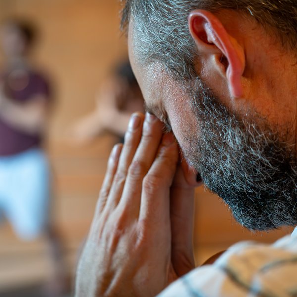 NEU: Yoga- & Ayurveda-Retreat Bärtiger Mann hält die Hände vor dem Gesicht, Person im Hintergrund