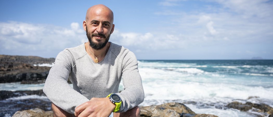 Mann mit Bart und grauem Pullover sitzt auf Felsen am Meer bei sonnigem Wetter