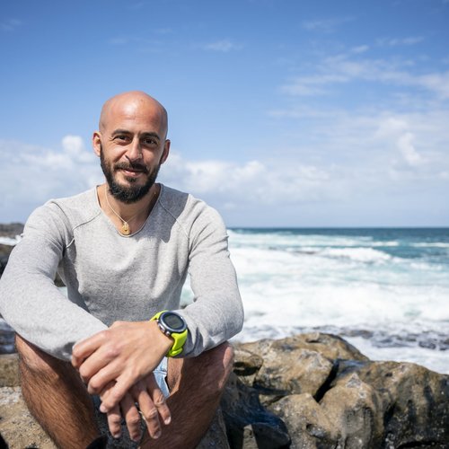 Mann mit Bart und grauem Pullover sitzt auf Felsen am Meer bei sonnigem Wetter