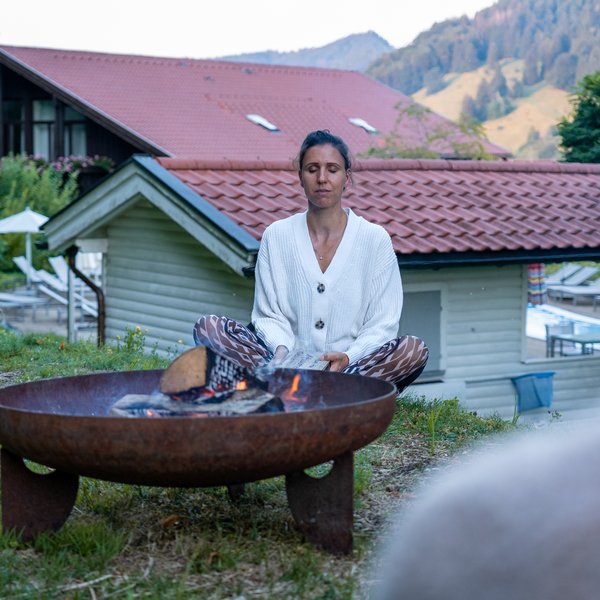 NEU: Yoga- & Ayurveda-Retreat Meditierende Frau neben einer Feuerschale vor einem Holzhaus in den Bergen