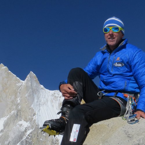 Bergsteiger in blauer Jacke auf Felsen mit schneebedecktem Berg und klarem Himmel
