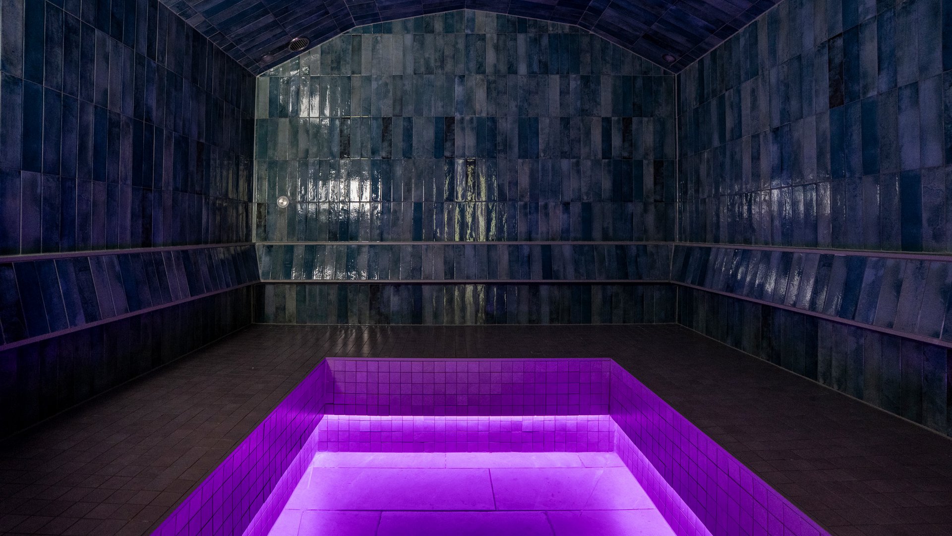 Dunkler Raum mit glänzenden blauen Fliesen und lila beleuchtetem Becken in der Mitte