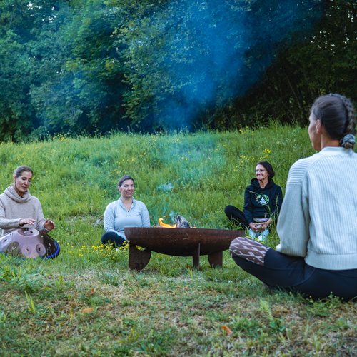NEU: Yoga- & Ayurveda-Retreat Vier Frauen sitzen auf einer Wiese um eine Feuerschale im Wald
