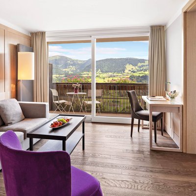 Hotel im Allgäu: 5-Sterne-Gesundheitsurlaub in Bayern Helles Wohnzimmer mit Sofa, lila Sessel, Schreibtisch und Balkon mit Bergblick