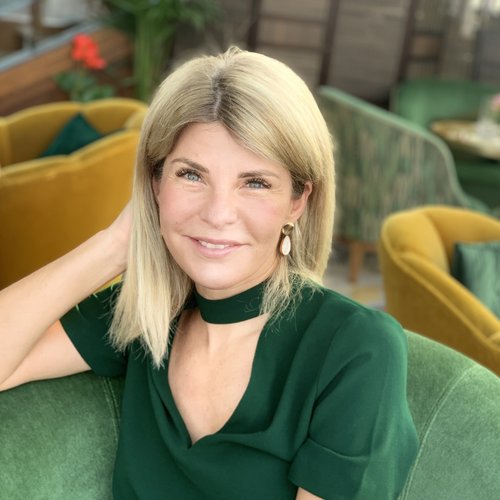 Wissen, das inspiriert: Unsere Vortragsreihe greift Themen auf, die das Leben bereichern. Frau mit blondem Haar und grünem Kleid sitzt und lächelt in einem Café