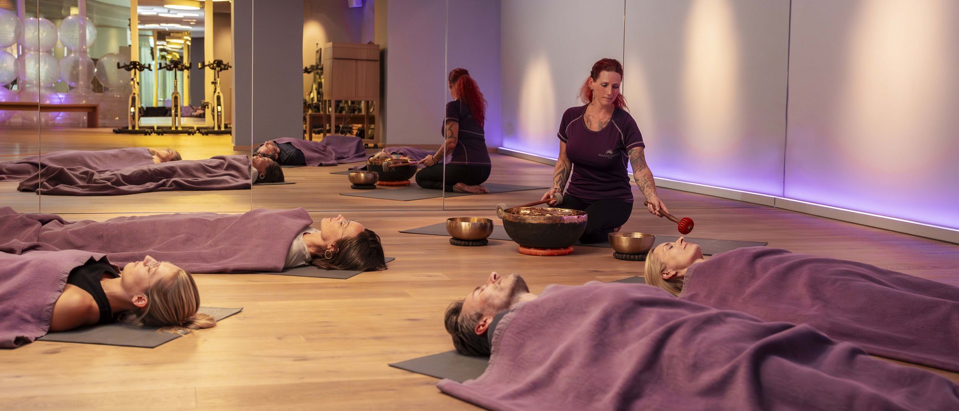 People lying covered with blankets as a woman plays singing bowls in a room