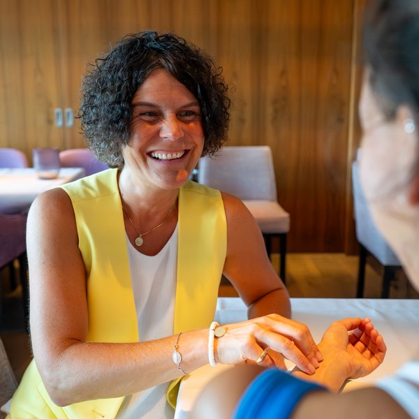 NEU: Yoga- & Ayurveda-Retreat Lächelnde Frau in gelber Weste im Gespräch mit einer Person im Restaurant